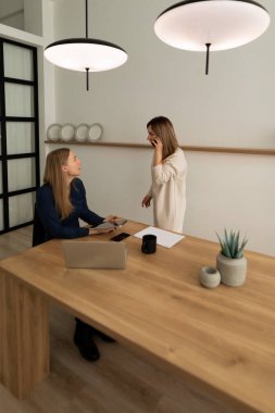 Çağdaş bir ofis ortamında iki kadın konuşuyor. Biri dizüstü bilgisayar ve not defteriyle ahşap bir masada otururken, diğeri de yakınlarda bir telefon görüşmesi yapıyor..