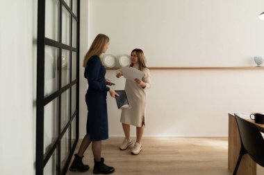 İki kadın, minimalist bir ofis ortamında ortak bir toplantıya katılıyor. Zarif bir duvar dekorunun yanında dururken belge ve fikir alışverişinde bulunurlar..