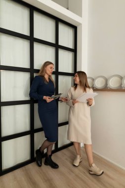 İki kadın ellerinde belgelerle bir sohbete girişiyor. Minimalist dekor ve zevkli bulaşık takımlarıyla çevrili parlak siyah beyaz bir bölmenin karşısında duruyorlar..