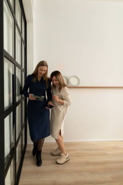 İki kadın şık, kapalı mekan ortamında birlikte, gülümsüyor ve dijital cihazlarla meşgul. Fikirlerini ve kahkahalarını paylaşıyorlar, dostça bir ortamda işbirliği gösteriyorlar..