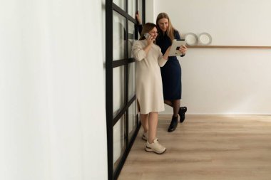 İki kadın ferah ve minimalist bir odada verimli bir konuşma yapıyorlar. Biri telefonu tutarken, diğeri tabletteki içeriği inceliyor. Doğal ışık alanı dolduruyor..