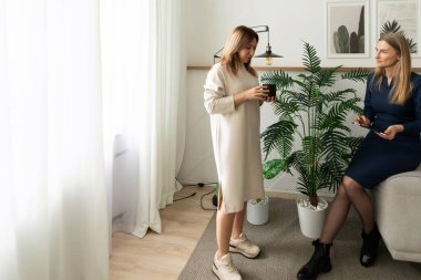 İki kadın, kapalı mekan bitkileriyle süslenmiş rahat bir odada ayakta ve otururken sohbet ediyorlar. Yumuşak doğal ışık perdelerin arasından süzülüp sıcak bir atmosfer yaratır..