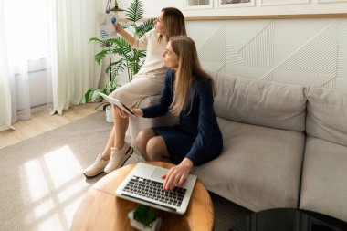 İki kadın şık, güneşli bir oturma odasında ortak bir tartışmaya giriyorlar. Biri duvardaki bir belgeyi işaret ediyor, diğeri ise içerideki bitkilerle çevrili bir dizüstü bilgisayardan notlar alıyor..