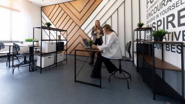 Çağdaş bir ofiste, bir kadın dizüstü bilgisayar kullanarak masada otururken, diğeri de yakınlarda durup fikirleri tartışıyor. Şık ortam yaratıcılığı ve işbirliğini teşvik ediyor..