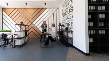 İki kadın, duvarda motivasyon ifadeleri ve çağdaş bir estetik tasarım içeren şık bir ofis alanında tartışmaya girişirler..