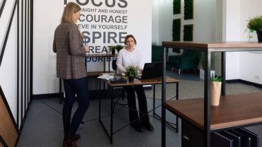 Şık bir ofis ortamında, iki kadın masa başında dikkatle sohbet eder. Biri yeşil ve motivasyonel ifadelerle çevrili bir dizüstü bilgisayarla otururken diğeri ayakta duruyor..