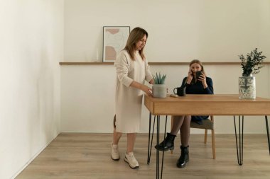 Bir kadın bir bitkiyle ilgilenen tahta bir masanın başında otururken, diğeri de yanında oturup dikkatlice dinliyor. Uzay, sakin bir atmosfer yaratan temiz çizgiler ve yumuşak renklere sahiptir..