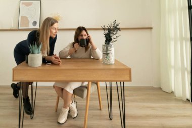 İki kadın, modern ve parlak bir çalışma alanında yaratıcı bir an yaşıyor. Bir kadın fotoğraf çekerken, diğeri dikkatlice izliyor. Etrafı bitkiler ve zarif dekorlarla çevrili..