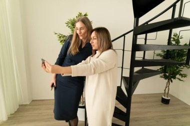 İki kadın birlikte poz veriyor, gülümsüyor ve selfie çekmek için akıllı telefon tutuyor. Çevrelerinde yeşillik olan çağdaş bir kapalı alandalar..