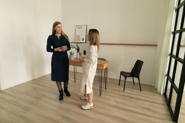 İki kadın parlak bir ofiste önemli bir şeyi tartışıyor. Biri mavi bir elbise giyerken, diğeri açık renkli bir elbise giyiyor. Ahşap bir masa ve sandalye ayarları tamamlar..