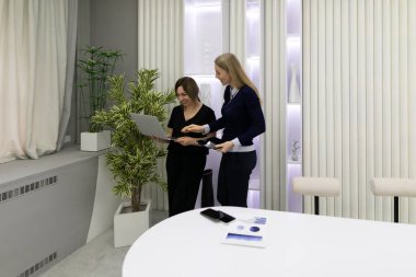 İki kadın şık bir ofiste dizüstü bilgisayarı incelerken bir tartışma içindeler. Parlak, modern tasarım elementleri ve bitkiler takım çalışması ve yaratıcılık için davetkar bir atmosfer yaratır..