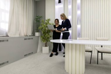 Çağdaş bir ofiste yan yana duran iki kadın dizüstü bilgisayarı inceliyor ve proje detaylarını etraflarında şık bir masa ve kapalı mekan bitkileriyle tartışıyorlar..