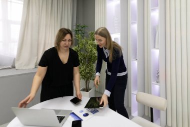 Çağdaş bir ofiste birlikte çalışan iki kadın, fikirleri tartışıyor ve beyaz bir masa üzerinde birden fazla cihaz kullanırken, etraflarında parlak dekor ve yeşillik var..