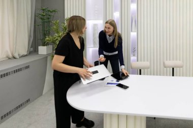 İyi ışıklandırılmış modern bir ofiste, iki kadın ortak bir tartışmaya girişirler. Bir kadın dizüstü bilgisayarla dururken, diğeri masadaki belgeleri işaret ediyor..