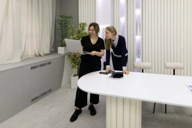 İki kadın şık bir ofis ortamında dizüstü bilgisayara bakarken bir tartışmaya girişirler. Oda modern dekor, hafif tonlar ve beyaz bir masaya sahiptir..