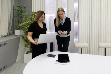 Modern bir ofis alanında iki kadın iş birliği yapıyor. Bir dizüstü bilgisayar ve akıllı telefondan dokümanları inceliyorlar. Etrafı bitkiler ve çağdaş dekorlarla çevrili gösterişli bir masanın yanında duruyorlar..