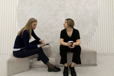 İki kadın şık ve minimalist bir ortamda oturup sohbet ediyorlar. Bir kadın elinde kahve fincanı tutarken tablet kullanıyor. Etkileyici etkileşimlerini vurguluyor..