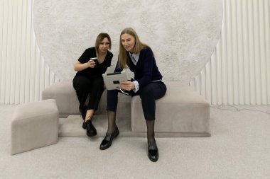 İki kadın dijital bir cihaz kullanırken işle ilgili bir tartışmaya giriyorlar. Modern, minimalist bir ortamda şık bir koltukta oturuyorlar..