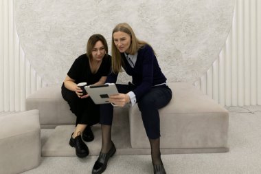 İki kadın modern bir kanepede birlikte oturuyor, biri kahve fincanı tutuyor. Bir tablet cihazına odaklanırlar, profesyonel bir çalışma alanında bilgi tartışırlar..