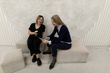 İki kadın şık bir iç mekanda oturmuş, canlı sohbetler yapıyorlar. Biri bir içki içerken diğeri de rahatlamış bir atmosferde gülümseyen bir cihazdan bir şey paylaşıyor..