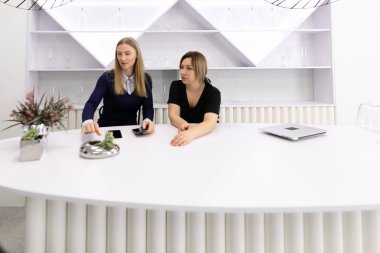 Minimalist bir ofiste şık bir yuvarlak masada sohbet eden iki kadın. İşle ilgili konuları tartışırken dekoratif bir öğeyi analiz ediyorlar.