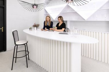 İki kadın beyaz, tırtıklı bir bar tezgahında oturuyorlar, dijital cihazlar kullanarak sohbet ediyorlar. Parlak çalışma alanı şık dekorlar ve modern ışıklandırmalar içerir..