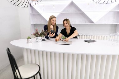 İki kadın gülümser ve minimalist dekorlarla çevrili parlak beyaz bir masada fikirlerini paylaşırlar. Sıcak ve profesyonel bir atmosfer sergileyen bir projede işbirliği yapıyorlar..