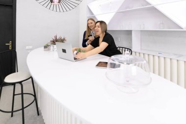 İki kadın, doğal ışıkla dolu çağdaş bir çalışma alanındaki şık beyaz bir tezgaha yerleştirilmiş bir dizüstü bilgisayar üzerinde ortak bir tartışmaya giriştiler, temiz bir estetik sergiliyorlar..