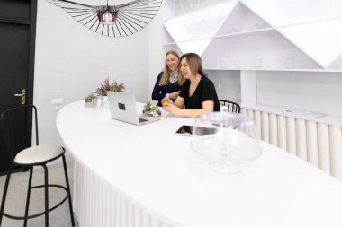 Çağdaş bir ofis ortamında, iki kadın dizüstü bilgisayarları üzerinde çalışırken sohbet ediyorlar. Minimalist tasarım beyaz mobilyalar ve şık dekorlar içerir..