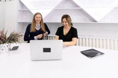 İki kadın minimalist bir çalışma alanında parlak bir masada oturmuş dizüstü bilgisayara bakarken fikirleri tartışıyorlar. Profesyonel bir ortamda işbirliği ve yaratıcılığı gösteren bir not defteri var..