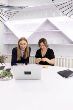 İki kadın modern beyaz bir masada şık bir ofiste, ortak bir çalışma oturumunda oturuyor. Fikirleri tartışırken ve laptopta bilgi incelerken atıştırmalık yemek yemek hoşunuza gider..