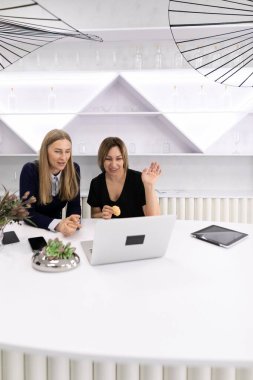 İki kadın, çağdaş bir ofiste, gösterişli beyaz bir masada ortak bir tartışma içindeler. Atıştırmalıkları paylaşıyorlar ve bir dizüstü bilgisayarla etkileşime geçiyorlar, takım çalışmasını ve yaratıcılığını teşvik ediyorlar..