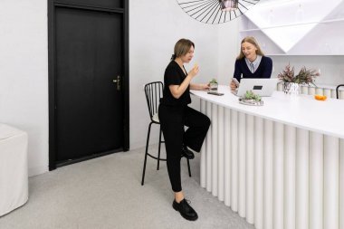 Çağdaş bir mutfakta, bir kadın bir taburede oturmuş laptopta çalışan başka bir kadınla sohbet ediyor. Rahat bir anı paylaşıyorlar..