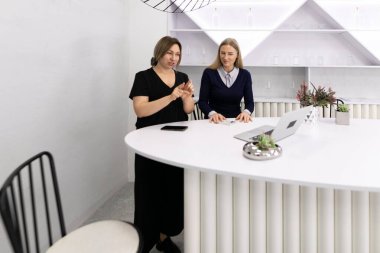 Çağdaş bir ofiste şık beyaz bir masada sohbet eden iki kadın. Fikirlerini paylaştıkça bir jest, temiz çizgiler ve minimum dekorla çevrili, işbirlikçi bir çevreyi işaret ediyor..