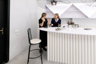 Şık bir ofis alanında, iki kadın çağdaş bir resepsiyon masasında canlı canlı sohbet ediyorlar. Parlak ışıklandırma temiz tasarımı geliştirir, profesyonel bir atmosferi gösterir..