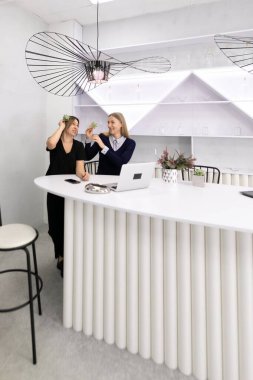 İki kadın, çiçeklerle süslenmiş şık bir resepsiyon masasında eğlenceli bir etkileşimi paylaşıyor. Modern alan parlak ve davetkar, çağdaş tasarım elementleri içeriyor..