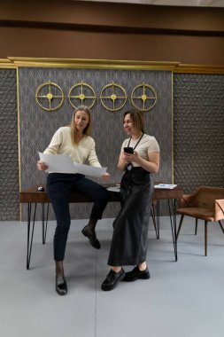 İki kadın şık bir ofis alanında tasarım planlarını tartışıyor. Bir kadın masada otururken diğeri yakınlarda oturup sohbet ediyor. İkisi de odaklanmış ve yaratıcı görünüyorlar..