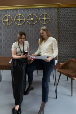 Çağdaş bir ofiste iki kadın aynı anı paylaşıyor. Biri masada otururken diğeri ayakta duruyor, ikisi de gülümsüyor ve akıllı telefona bakıyor. Parlak ve şık bir ortam havayı yükseltir..