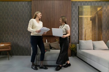 İki kadın, şık bir ofis alanında proje belgeleriyle ilgili bir sohbete girişiyor. Atmosfer, rahat mobilyalar ve zarif dekorlarla birlikte işbirlikçi ve profesyoneldir..