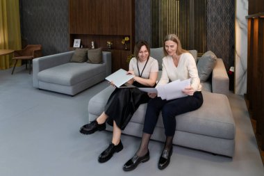 İki kadın şık bir salonda rahat bir koltukta oturmuş proje belgelerini gözden geçiriyorlar ve birlikte çalışırken fikirleri tartışıyorlar..