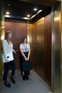 İki kadın, ahşap aksanlı modern bir asansörde ellerinde iş eşyalarıyla bir şeyleri tartışıyorlar. Bu ortam tipik bir şirket ortamı..