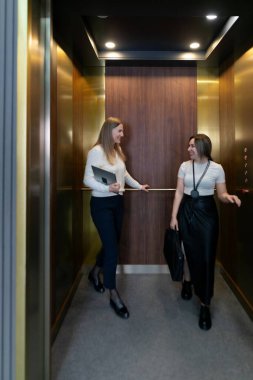 İki kadın asansörde canlı bir sohbet yapıyorlar. Biri bir tablet tutuyor, diğeri bir çanta taşıyor. İçerisi parlak ve çağdaş, sıcak ışıklandırmalı..
