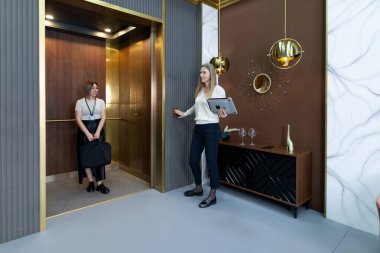 İki profesyonel şık bir asansörün içinde sohbet ediyorlar. Bir kişi tablet tutarken, diğeri küçük bir çantayla ayakta duruyor. Uzay, çağdaş tasarım elementlerini içerir.