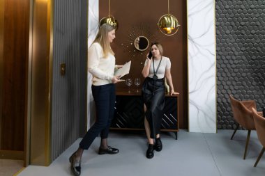 İki profesyonel kadın şık bir ofis ortamında sohbet ediyor, biri bir klasör tutarken diğeri de telefonuyla konuşuyor, iş yerinde ortak bir an sergiliyor..