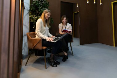 İki kadın şık bir çalışma alanında otururken verimli bir tartışmaya girişirler. Biri bir dizüstü bilgisayarı incelerken, diğeri yakınlarda çağdaş dekorlarla çevrili bir kitap okuyor..