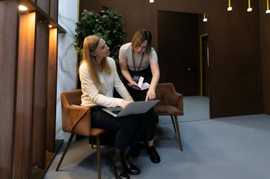 İki kadın, çağdaş bir ofis ortamında dizüstü bilgisayar üzerinde çalışırken profesyonel bir tartışmayla meşgul oluyor. Çevrede zarif mobilyalar ve bitkiler var..