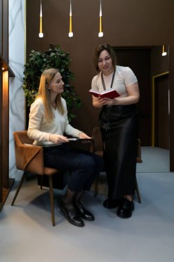 Bir kadın elinde bir bloknotla otururken diğeri ayakta duruyor ve çağdaş ve zevkli bir şekilde tasarlanmış bir iç mekanda dizüstü bilgisayardan bilgi sunuyor..