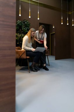 İki kadın modern bir ofiste birlikte oturup, minimalist dekorlar ve yumuşak ışıklandırma ile çevrili bir belge üzerinde tartışırken ve çalışırken.