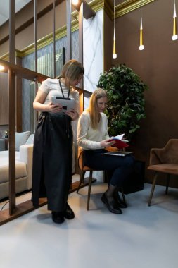 İki kadın şık bir ofis ortamında birlikte çalışırlar. Biri tablet tutarken diğeri ise güncel mobilyalarla çevrili belgelerle oturuyor..