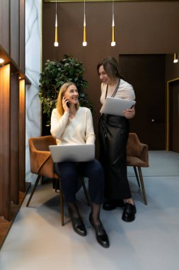 Şık bir ofis ortamında, biri telefonda konuşurken iki kadın birbiriyle etkileşir. Çağdaş dekorlar, dizüstü bilgisayarlar ve yeşilliklerle çevrili olup, işbirlikçi bir atmosferi vurguluyorlar..